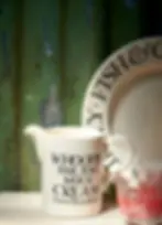 The Gift Guide for Home Lovers 2025: Festive Fridays – Earn Rewards While You Shop. Cream jug with humorous text, "WHOOPS! FAR TOO MUCH CREAM (but how DELICIOUS)", sits by a decorative plate and glasses against a rustic wall.