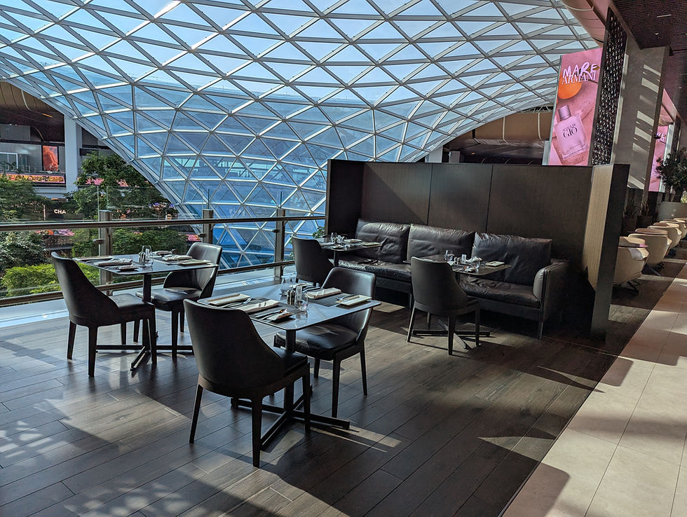 Modern interiros of Al Mourjan Garden Lounge with black leather seating, set tables, and a glass-paneled wall. A geometric ceiling and pink advertisement in the background.