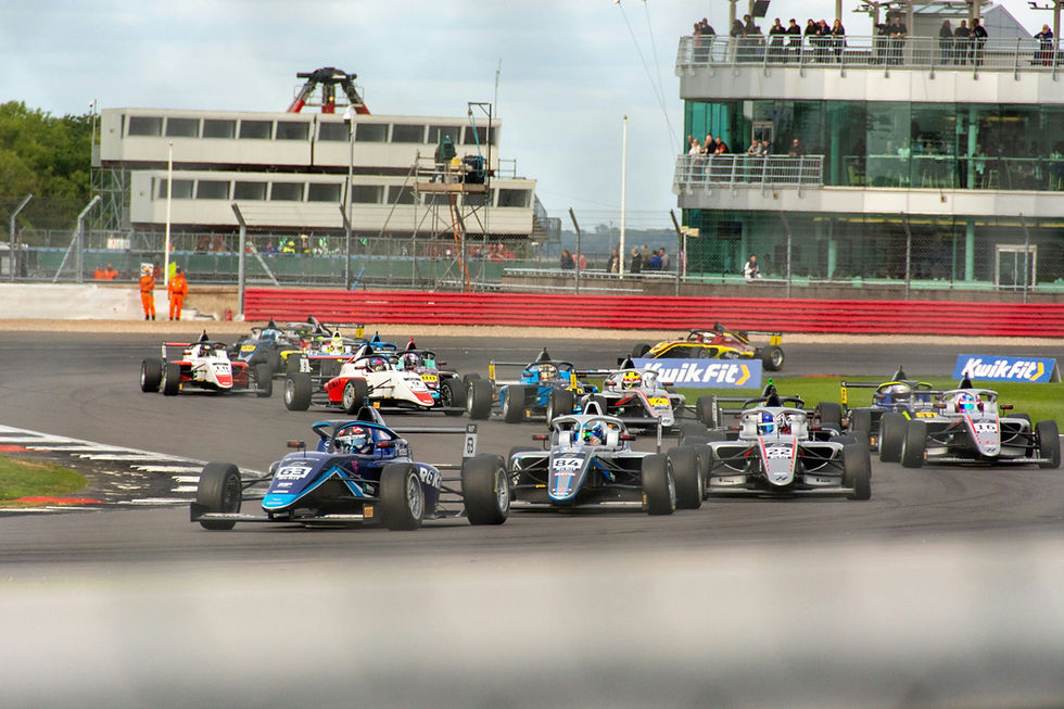 Last Chance for Amex Cardholders: Secure Your Silverstone F1 2026 Presale Tickets Now. Race cars speed on a track with spectators in a glass building. Bright race suits and "Kwik Fit" signs are visible. Energetic atmosphere.