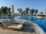 Poolside loungers with striped cushions and closed umbrellas by a calm blue pool. Trees and mountains in the background, serene setting.