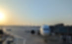 Enjoy up to 60% More Avios with Qatar Airways’ Limited-Time Offer. Plane at airport gate during sunrise, with a clear sky. Tarmac vehicles include a green cart and a white tug labeled "DOUGLAS." City skyline in distance.