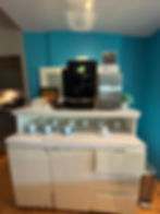 Coffee station with a machine on a white counter, surrounded by cups and saucers. Blue wall in background, plant and brochures visible.