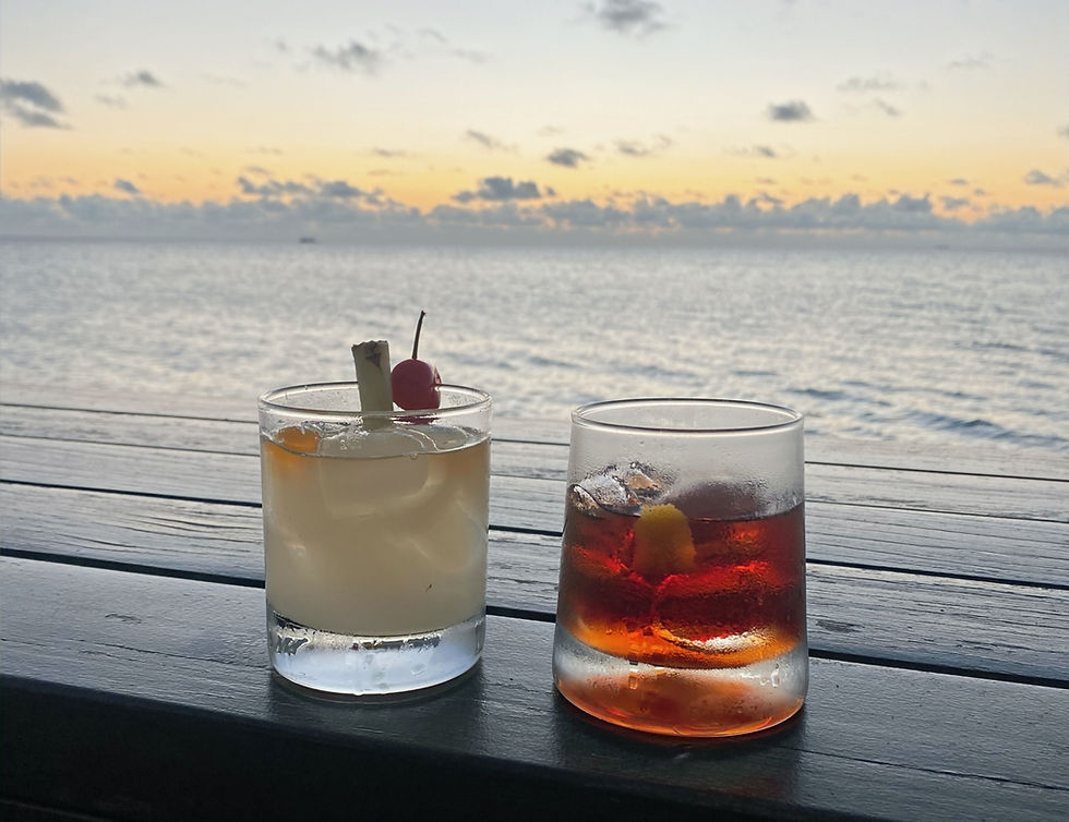Two cocktails on a wooden table by the ocean at sunset, with a calm, serene ambiance. One has a cherry garnish, the other an orange peel.