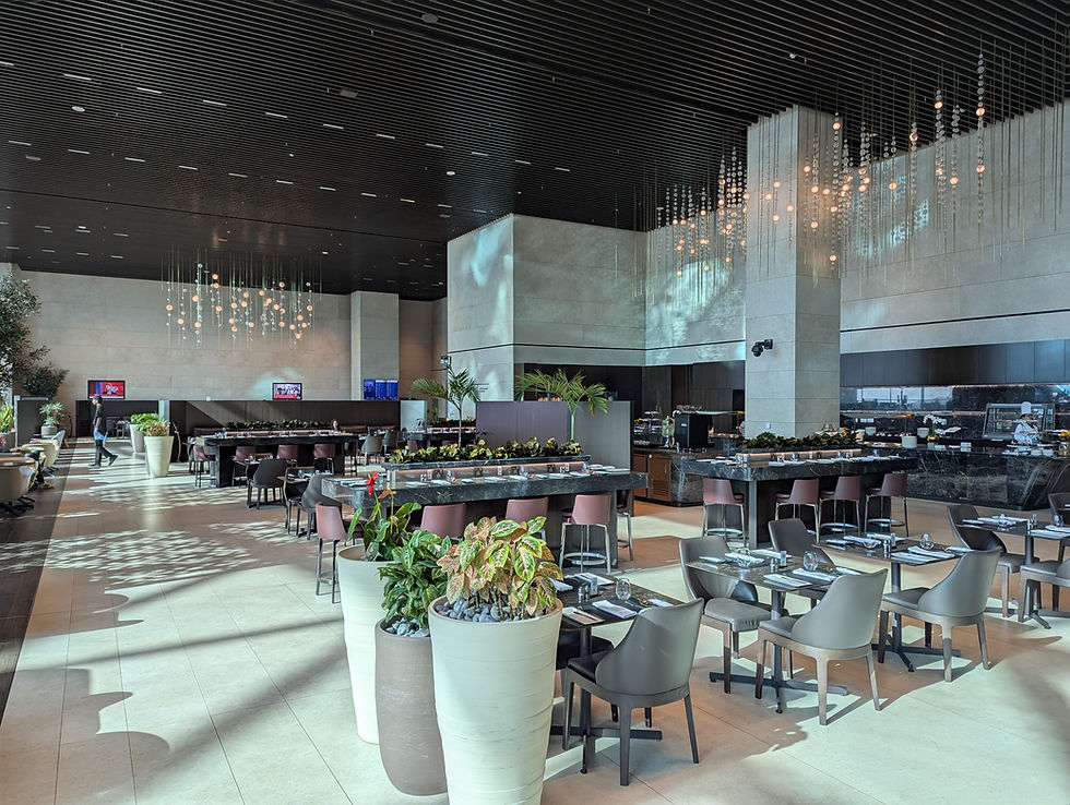 Spacious modern restaurant with high ceilings, pendant lights, and plants. Empty tables and chairs, two TVs on walls, and a sleek, elegant ambiance.