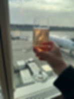 Qatar Airways A380 Business Class Review Heathrow to Doha. Hand holding champagne flute by airport window, airplane outside. Sunlit bubbles, relaxed travel mood. Sky and tarmac in background.