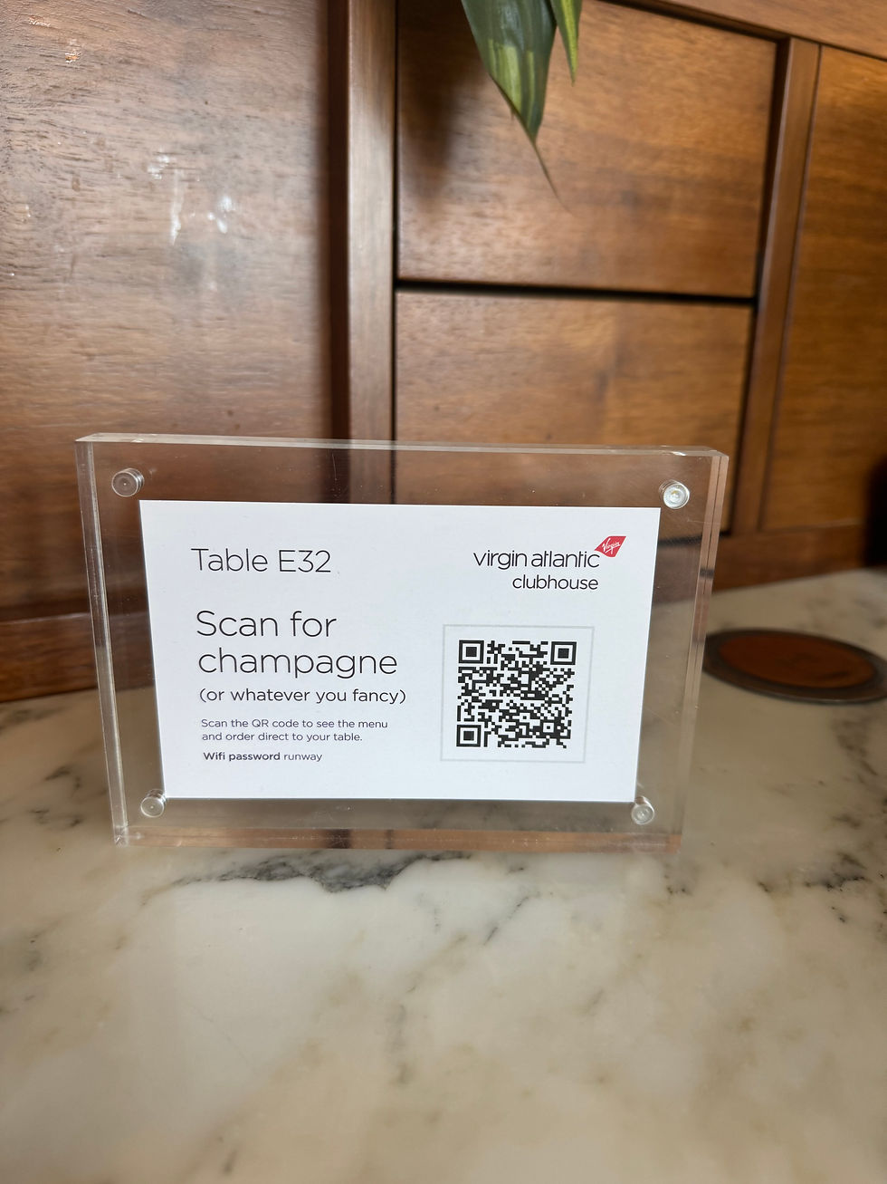 A table sign with a QR code for menu ordering at "Table E32" in Virgin Atlantic Clubhouse, set on a marble surface with wood backdrop.