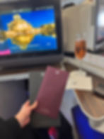 Qatar AirwaysA380 Business Class Review, Heathrow to Doha. Airplane seat with a screen displaying "Oryx One." A hand holds two menus. A glass of pink champagne sits on the tray table.