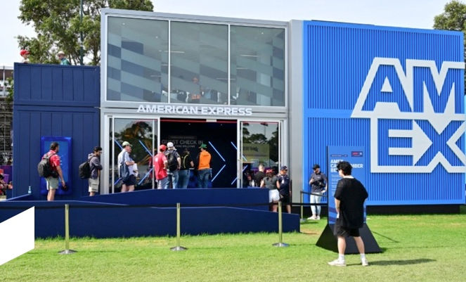 People queue outside a blue building labeled "American Express" in a grassy area. The mood is lively with a clear sky.