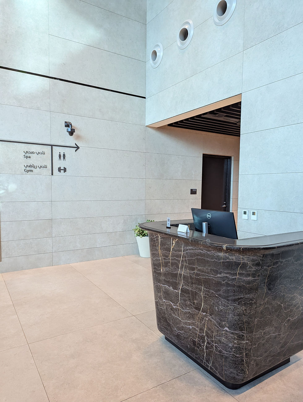 Reception area with directional signage to the spa and gym at Al Mourjan Garden Lounge in Doha Airport with a marble desk, potted plant, and signage for spa and gym. Beige walls, modern design, calm atmosphere.