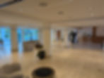 Spacious hotel lobby with beige sofas, potted plants, and large windows. A wooden reception desk is in the background. Bright, calm atmosphere.