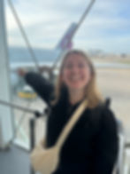 Qatar Airways A380 Business Class Review Heathrow to Doha. Smiling woman in black points at airplane outside terminal window. Clear sky, plane with maroon tail logo visible. Airfield in background.
