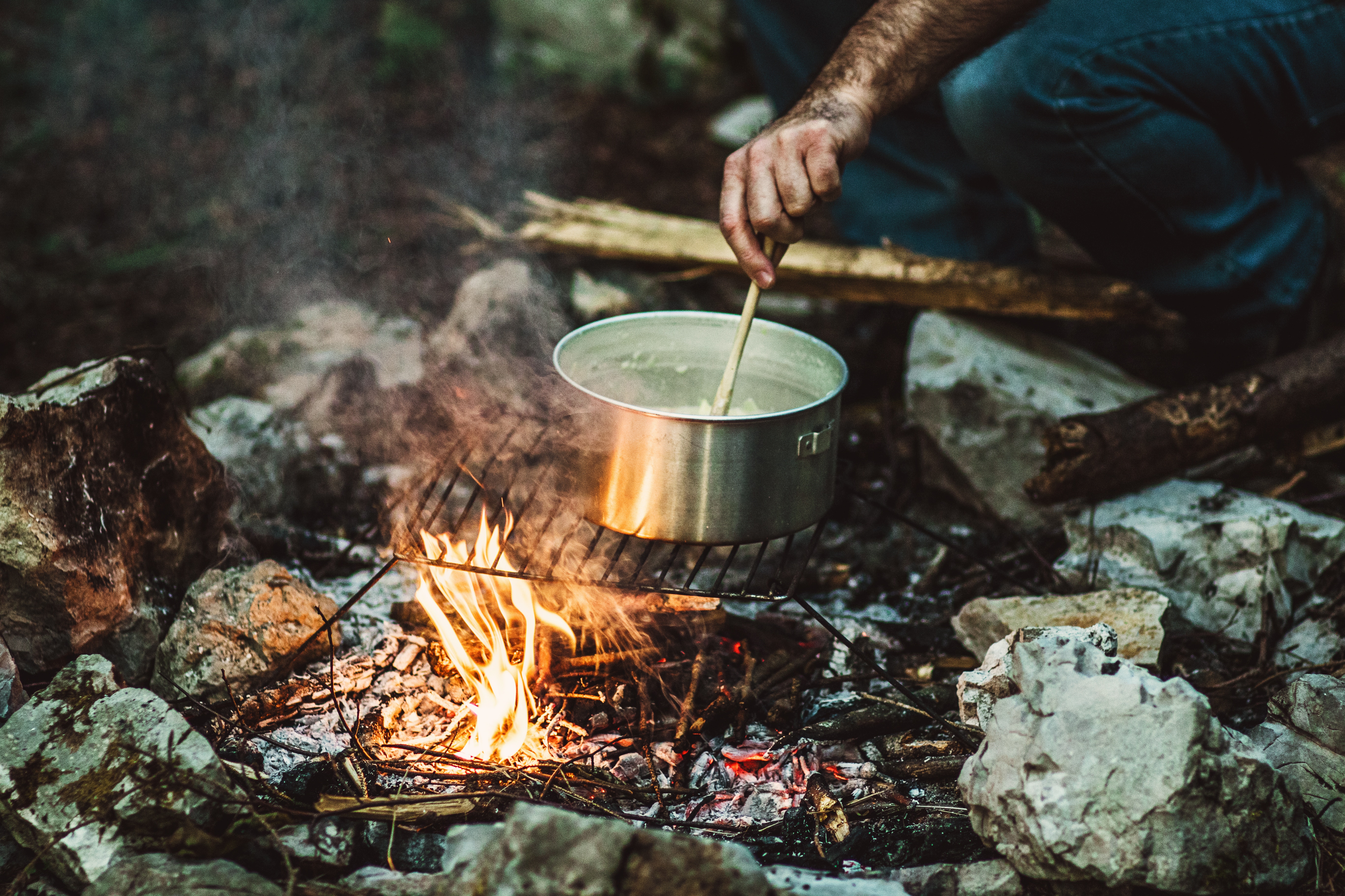 Man with muddy arm stirring pot of hot water and vegetables over fire-pit campfire 