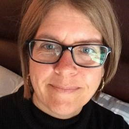 A portrait photo of our Trustee Dianne Beaumont smiling at the camera, wearing a black high neck shirt and solid black framed glasses, with shoulder height mousey brown hair.