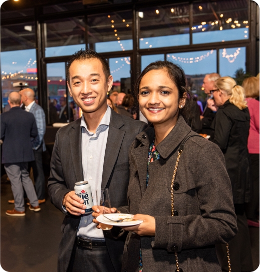 Two Cheers for Children attendees smiling, holding a beer can and food plate, with other guests and string lights in the background