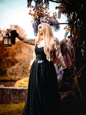 Tattoo-Model in einem schwarzen Kleid mit feinen Mustern, hält eine Laterne und steht unter blühendem Blauregen. Outfit mit Fairy-Vibes von Silvermoon Threads