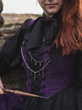 Detailaufnahme eines violetten Mieders mit Schnürung und filigranen Ketten, kombiniert mit schwarzer Bluse und Zauberstab, im witchy Dark Academia Stil.