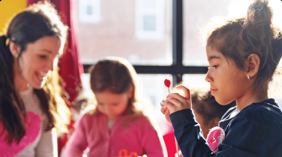 Young children engaged in a creative classroom activity with a teacher. A girl in the foreground focuses intently on a small red craft item, while other children and the teacher smile and work together in the background near a sunlit window.
