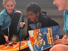 Three upper elementary students engaged in a drone activity, with one student holding a "Mission 4: Shark Watch" guidebook and another launching a drone from a landing pad.