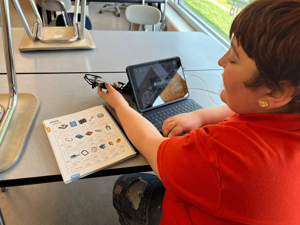 A student in a red shirt sits at a classroom table using a tablet to interact with a drone flight simulator. In front of them is a printed guide showing various drone components and controls. Sunlight streams through large windows in the background.