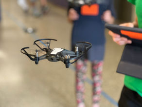 Close-up of a mini drone flying indoors, with students in the background holding tablets during a classroom STEM activity focused on safety and character development.