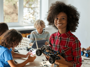 Smiling girl in a plaid shirt holding a wheeled robotics project in a STEM classroom with other students working on tech kits in the background.