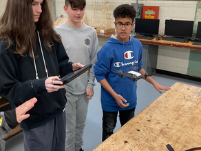 Three middle school boys operate a drone together in a classroom setting, using a tablet to navigate a flight mission as part of a team-based educational activity.