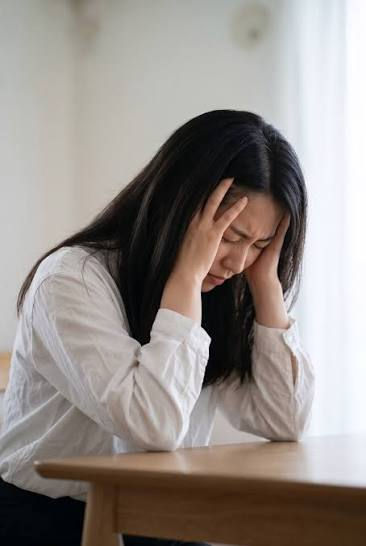 Distressed woman in a white shirt sits at a table, holding her head with eyes closed in a bright indoor room.