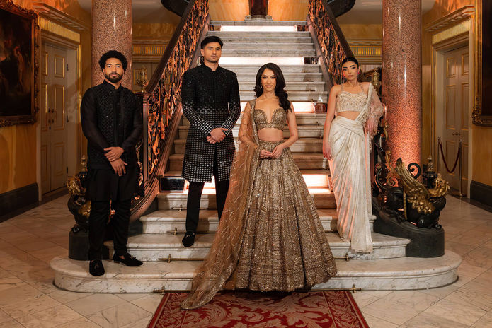 A group of four people in elegant traditional attire stand in front of a grand staircase. Two men wear dark sherwanis, and two women wear ornate lehengas. The setting is lavish, featuring marble and decorative columns.