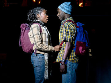 Two actors stare into each other's eyes. One holds onto the other's bag straps.