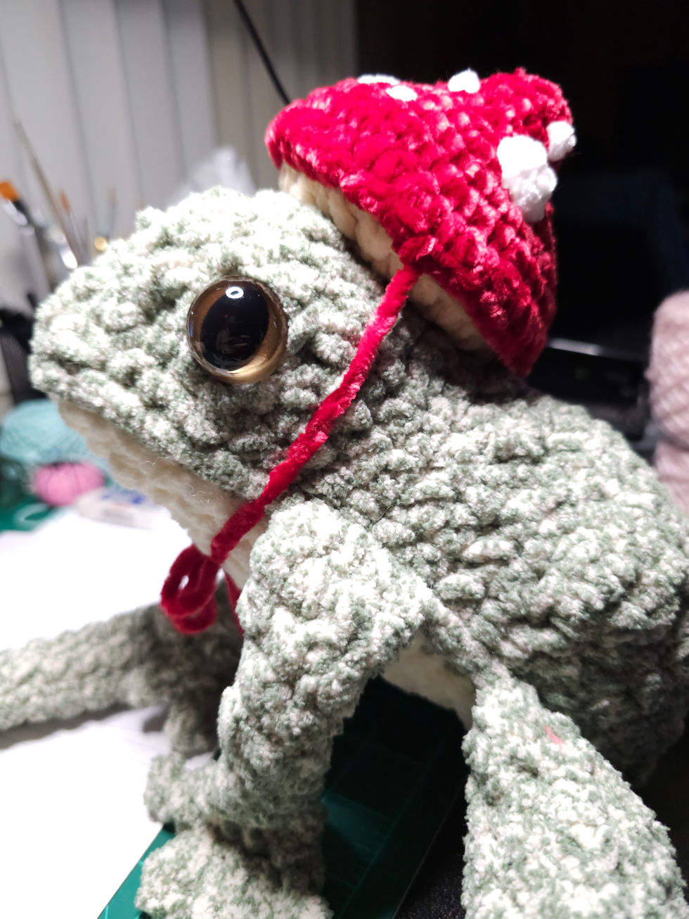 A large crocheted frog plushie wearing a red and white mushroom hat.
