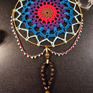 A dreamcatcher in bright blues that shift to purple and then to red. Beneath, there is a decorative string of wooden beads.