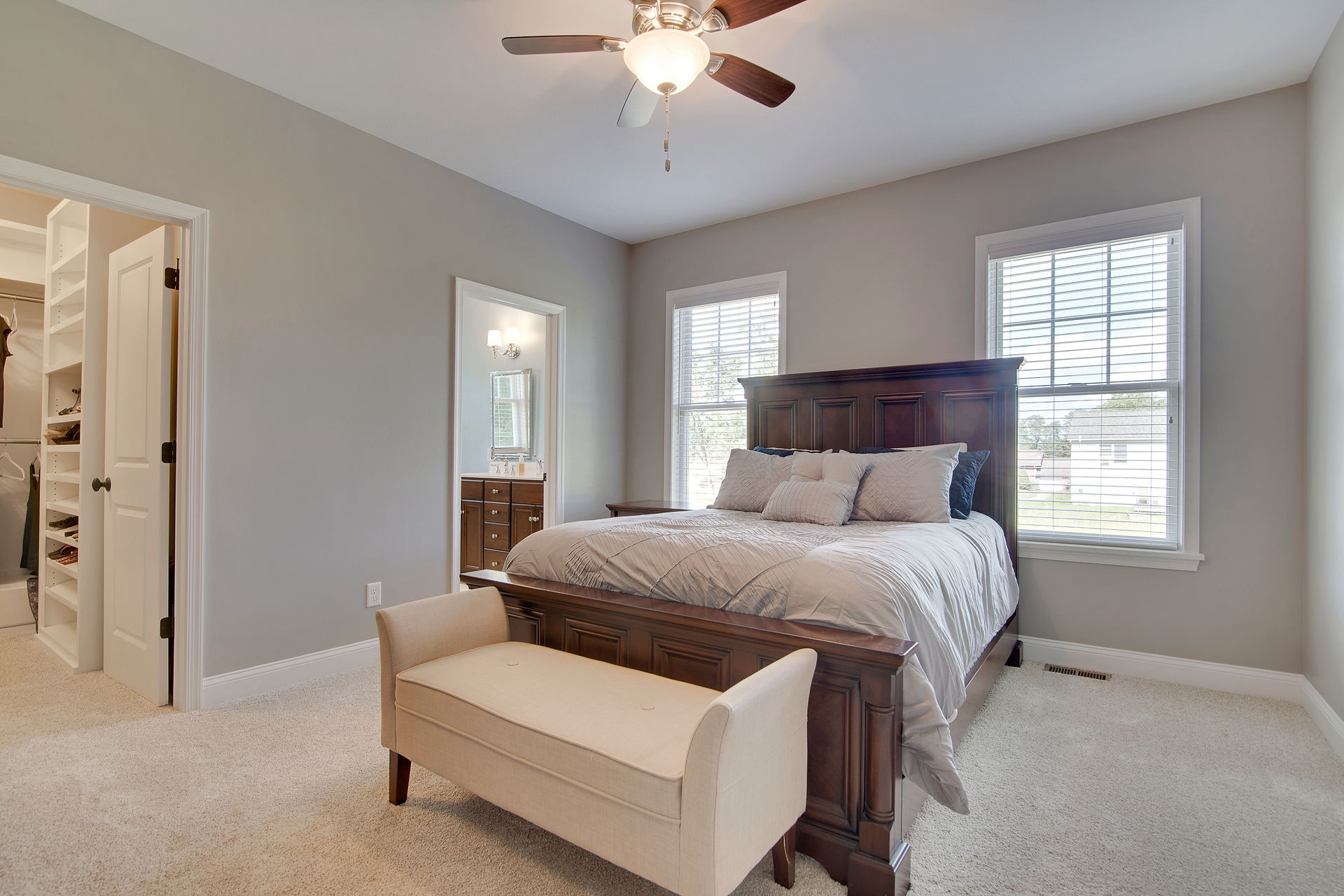 Furnished bedroom with beige carpet flooring and gray walls. The room has a large wooden bed frame with an paneled headboard, with light gray bedding and decorative pillows. A cream upholstered bench sits at the foot of the bed. Two windows with white blinds provide natural light.Open doorways lead to a walk-in closet with built-in shelving on the left and an en-suite bathroom with a dark wood vanity and wall-mounted lighting on the right. A ceiling fan with a light fixture is mounted overhead.