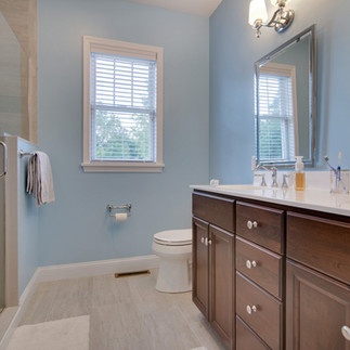 Finished bathroom with a double-sink vanity with dark wood cabinets and white countertops. Above the sinks are two framed mirrors and wall-mounted light fixtures. A glass-enclosed shower with tile walls is visible on the left, and a white toilet is beneath a window with white blinds. The walls are painted a light blue, and the flooring is light gray tile.