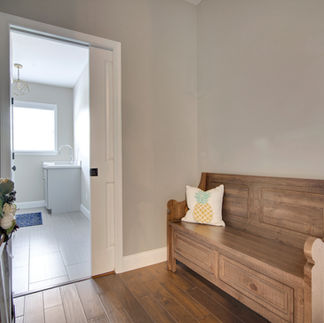 Mudroom hallway space with hardwood flooring leading into a laundry room with light gray tile flooring and a large window. A wooden bench with a decorative pineapple pillow sits against the right wall. On the left side of the hallway there is a wooden ladder with a floral arrangement. The laundry room has a built-in utility sink with a chrome faucet beneath the window, and a geometric pendant light hangs from the ceiling.