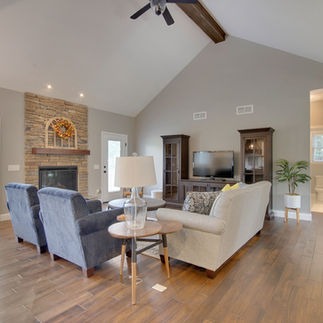 Furnished living room with vaulted ceilings and an exposed wood beam, with a central stone fireplace with a wood mantel and autumn wreath. The seating area has a light gray sofa, two navy blue armchairs, and a round wooden coffee table, and warm-toned hardwood flooring. A TV sits on a dark wood entertainment console with matching display cabinets. A side table with a glass lamp is placed next to the sofa, and there is a large potted plant in a white pot with wooden legs next to the doorway on the right.