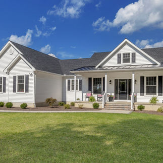 Exterior view of a single-story house in Stroud Township, PA with white siding, black shutters and a dark gray roof. The house has a wide covered front porch with two rocking chairs on the left side, a hanging swing on the right, and potted flowers along the steps. The lawn in front is expansive and neatly trimmed, with small shrubs and landscaped mulch beds lining the house.