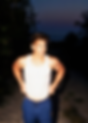 Waymon Hudson in a white tank top and blue shorts stands confidently on the beach at dusk, illuminated by warm twilight as the waves crash nearby. A snapshot of bold queer joy and sun-kissed self-expression in Costa Rica.