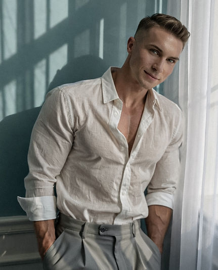 Stylish portrait of Waymon Hudson in an open white linen shirt and high-waisted trousers, leaning beside a softly lit window with a calm, confident expression. Editorial lifestyle image with warm neutral tones and cinematic lighting.