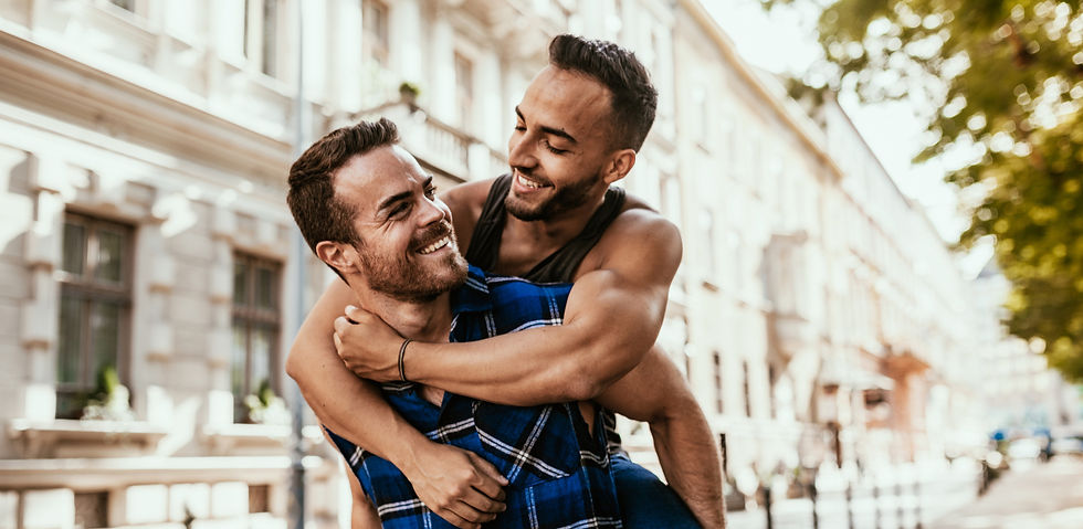 Gay couple smiling as one partner hugs the other from behind on a city street.