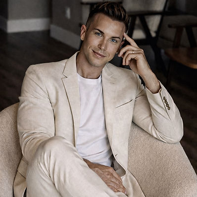 Waymon Hudson seated, wearing a cream blazer over a white t-shirt, one hand resting near his temple, looking at the camera.
