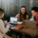 Three people laughing and working on laptops together inside a modern café.