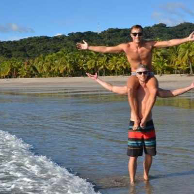 Two men at the beach, one lifting the other on his shoulders near the shoreline.