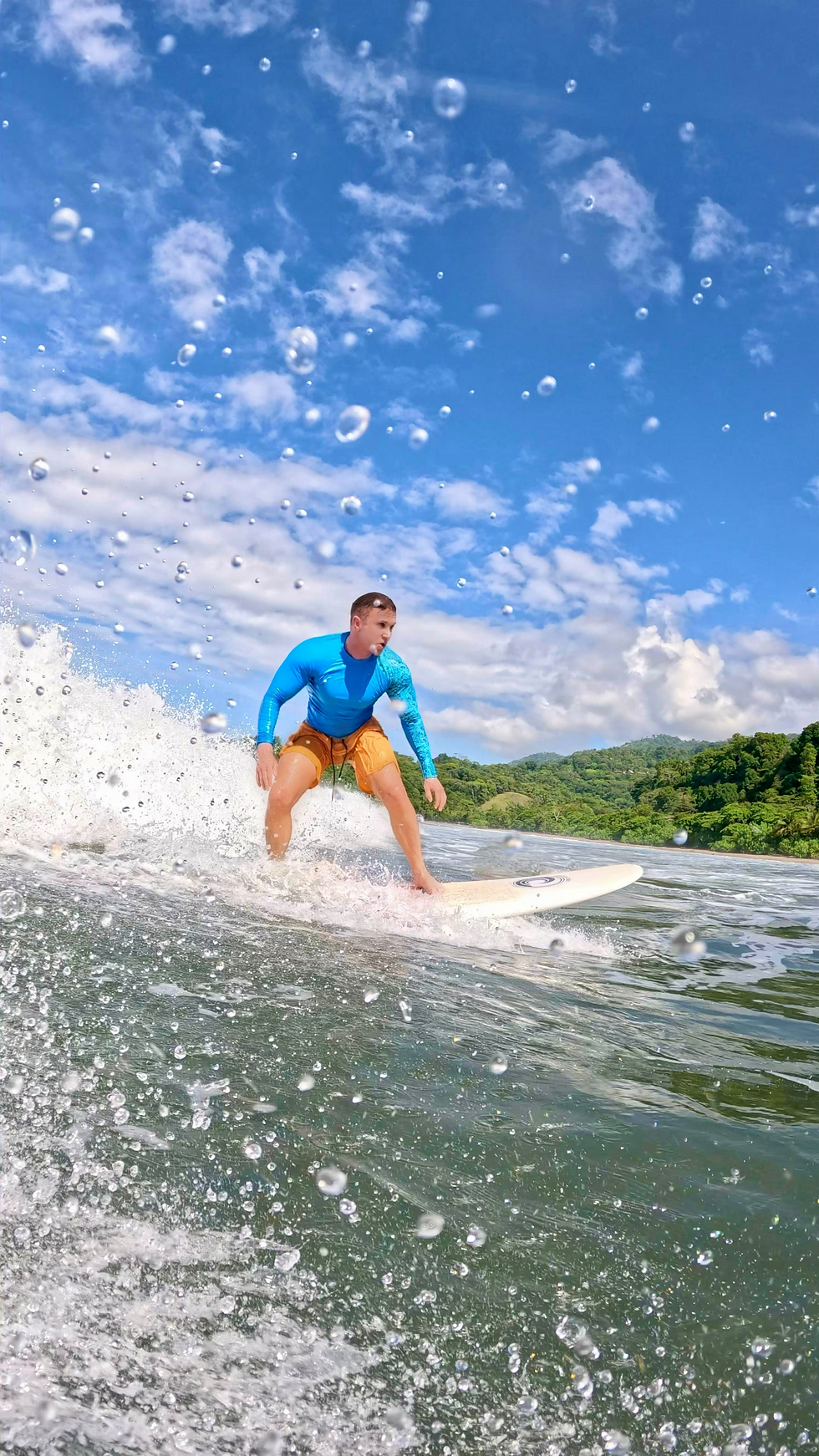 Waymon Hudson surfing in Costa Rica, riding a wave in a bright blue rash guard and orange board shorts, with jungle coastline and blue sky in the background — queer expat lifestyle