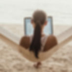 Woman working on a laptop while lying in a hammock on a sandy beach.