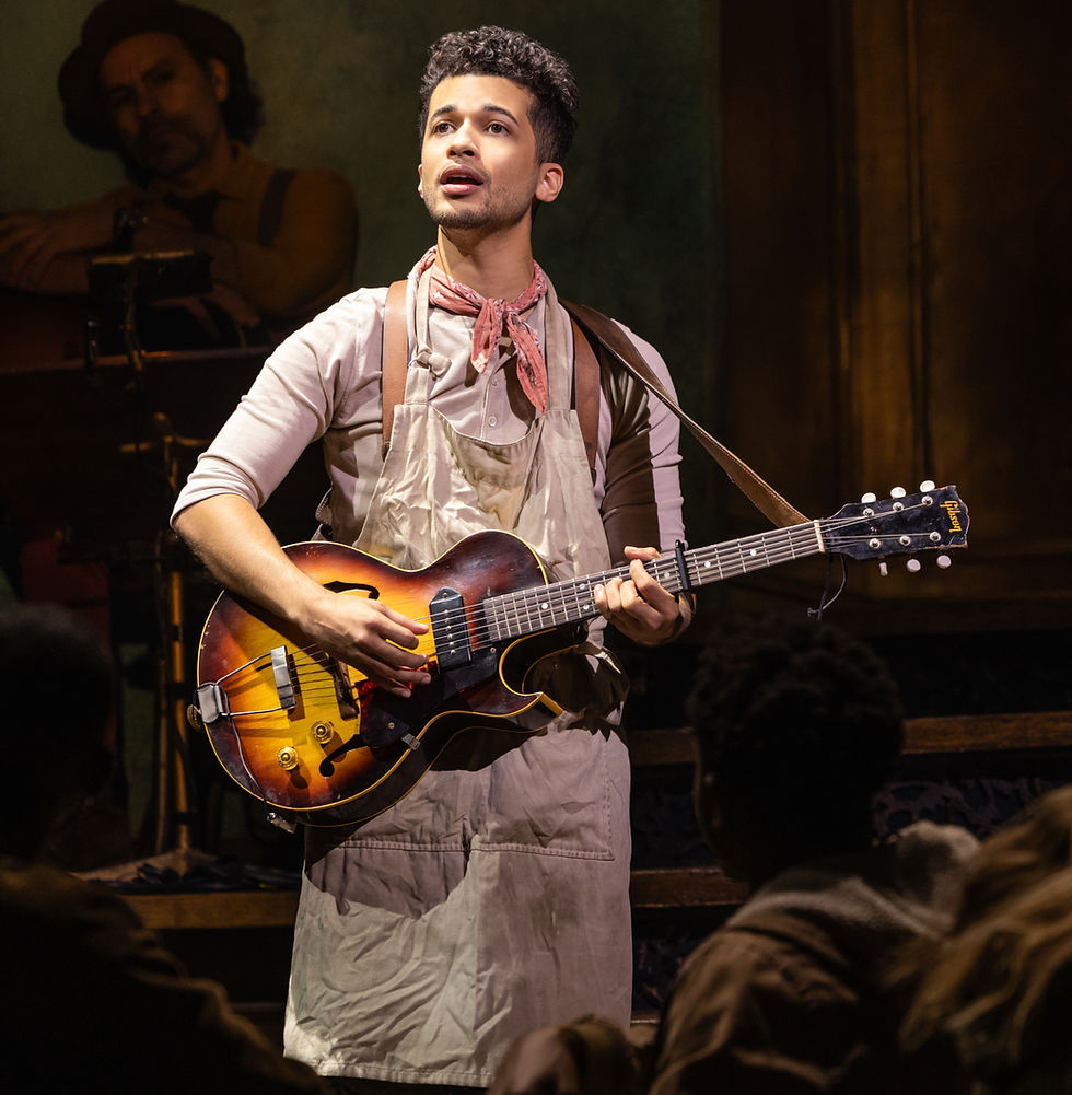 Jordan Fisher as Orpheus in Hadestown, standing center stage with a guitar, wearing a pale apron and red scarf, bathed in warm golden light — a soft, poetic figure in a hard, mythic world.