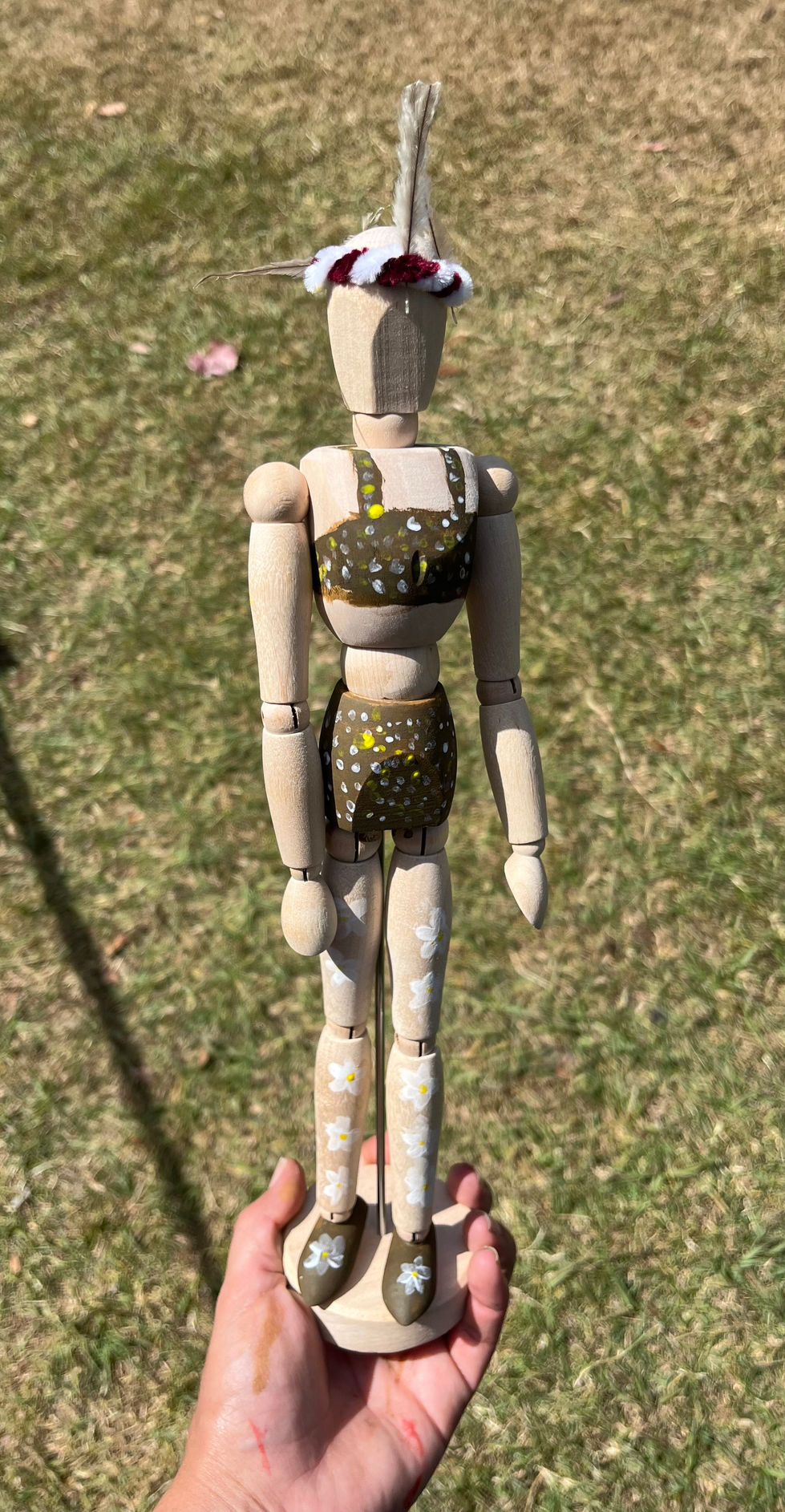 Wooden manikin doll wearing custom clothes, held in front of a yard.