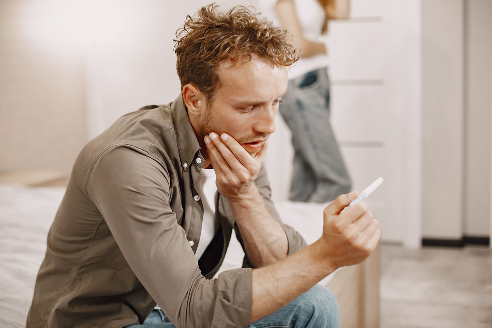 Worried man in a bedroom studies a pregnancy test, with a blurred woman in the background.