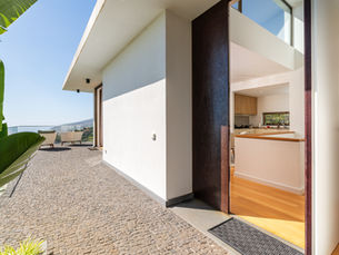 Entrada de uma moradia contemporânea na Madeira, com porta aberta, interior luminoso, terraço em pedra e vista ampla sobre o mar e a encosta.