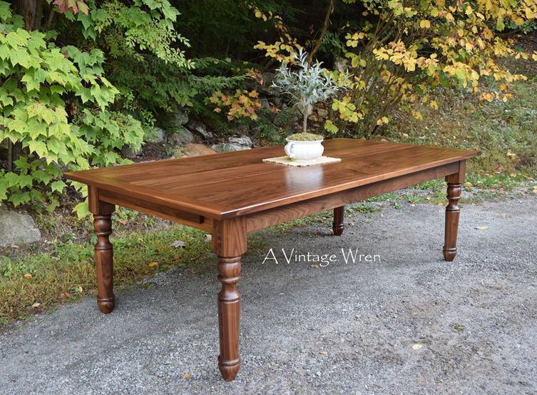 a New Hampshire made farmhouse dining table for 8 to 10. The table top is rich medium to dark brown colored wood made from solid Black Walnut.
The legs are turned on a lathe, they are chunky, large farmhouse style legs.
On the table top there is burlap runner with a mini olive tree in an antique ironstone pot.