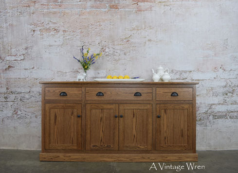 New Hampshire made solid wood sideboard buffet cabinet with four doors and three drawers. The color of the sideboard is a medium brown, the cup pulls and knobs are an oil rubbed bronze. The staged decor on the top of the sideboard includes a white porcelain cow creamer, vintage white ironstone tea set and a vase with yellow forsythia flowers
